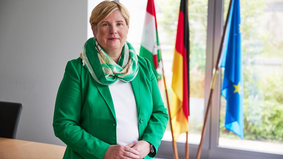 Claudia Middendorf sitzt an der Kante eines Tisches. Sie trägt eine dunkle Hose, eine weiße Bluse, einen grünen Blazer und einen farblich passenden Schal. Im hintergrund vor einem großen bodentiefen Fenster stehen die Flaggen des Landes Nordrhein-Westfalens, Deutschlands und Europas.