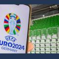 Auf dem Bild befinden sich im Hintergrund grüne Sitzplätze. Am linken Bildrand wird ein Handy in der Hand gehalten. Auf dem Display ist das Logo der Europameisterschaft abgebildet, ein Pokal auf unterschiedlichen Flaggen mit dem Schriftzug "UEFA EURO2024 GERMANY" darunter.