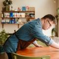 Ein junger Mann mit Down-Syndrom arbeitet in einem Café. Er trägt ein blaues Hemd und eine braune Schürze und wischt gerade einen Holztisch ab. Im Hintergrund steht ein Teil der Theke und es hängt ein Regal an der Wand. Darauf steht eine Pflanze  und verschiedene Packungen Kaffee.